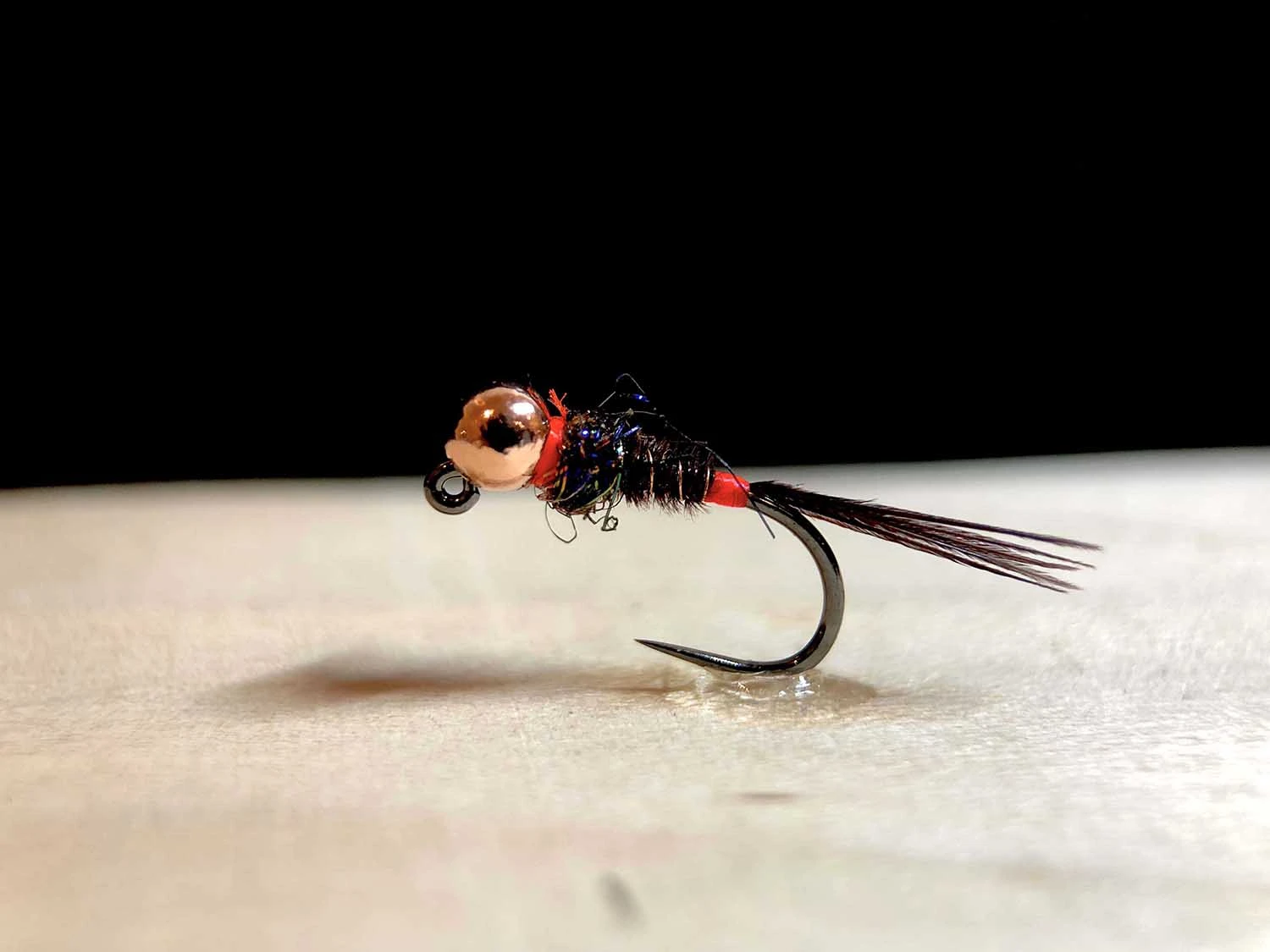 Beadhead Pheasant Tail nymph fly with copper bead and red hotspot on white surface